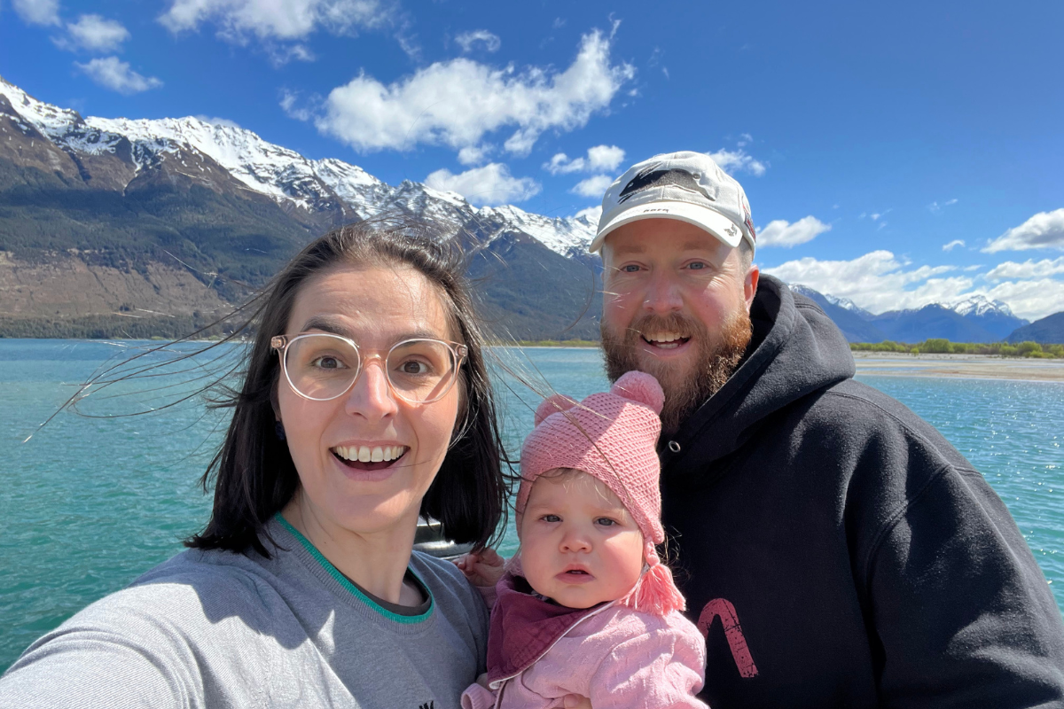 Dean with his partner and young child post on a holiday with ocean and snow-capped mountains behind them.